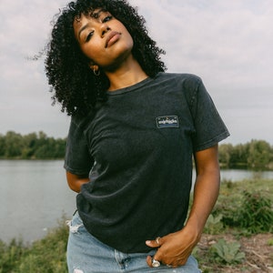 May include: A woman wearing a black t-shirt with a small rectangular patch that says "michigan" in yellow letters. She is wearing blue jeans with a distressed look and has curly black hair.