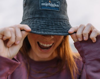 May include: A person wearing a black bucket hat with a white patch that says "EST. 1927 michigan" with a crescent moon and sun graphic. The person is wearing a purple long-sleeved shirt with a small patch that says "michigan" with a sun and wave graphic.