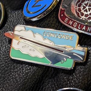 May include: A collection of enamel pins featuring various designs, including a plane with the text "CONCORDE", a skull with wings, and a rose with the text "ENGLAND".