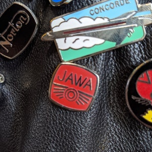 Può includere: Una giacca di pelle nera con varie toppe, tra cui una toppa "JAWA" rossa e bianca, una toppa "Norton" nera e bianca, una toppa "CONCORDE" blu e bianca e una toppa "FULHAM ANGLER CLUB" verde e oro.