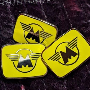 May include: Three yellow metal pins with a black and silver stylized letter M with wings on each pin.