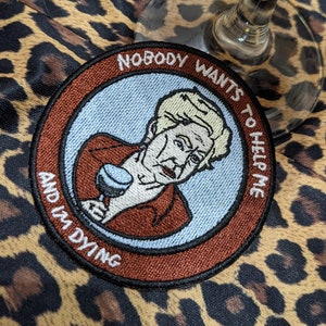 Könnte beinhalten: Ein runder gestickter Aufnäher mit hellblauem Hintergrund und braunem Rand. Das Motiv zeigt eine Person mit blonden Haaren, die ein Glas hält, umgeben von dem weißen Text: NOBODY WANTS TO HELP ME AND I'M DYING.