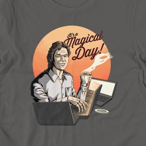 May include: A dark gray t-shirt with a cartoon illustration of a man sitting at a desk with a laptop and a cup of coffee. The text "It's a Magical Day!" is printed on the shirt.