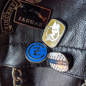 Puede incluir: Una chaqueta de cuero negra con varios parches, incluyendo un parche con el logotipo "Z" azul y blanco, un parche "Triumph" a cuadros blanco y negro, un parche "Lightning" dorado y blanco, un parche "Jaguar" negro con un pantera dorada y un parche "The Vincent" blanco y negro.