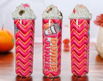 Op de afbeelding: Drie roze en oranje gestreepte bekers met witte deksels en een topping van slagroom. De bekers hebben het Dunkin'-logo en de tekst "Dunkie" erop gedrukt.