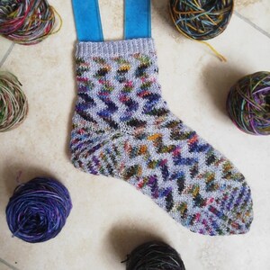 May include: A colorful sock with a geometric pattern, knitted with a variety of yarns in shades of blue, green, purple, and orange. The sock is displayed on a white tiled surface with several balls of yarn in various colors.