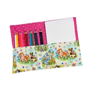 May include: A fabric pencil roll featuring a pink and white polka dot interior and a light-colored exterior with a pattern of kittens, flowers, and birdhouses. It contains eight colored pencils and a white notepad tucked into the pockets.