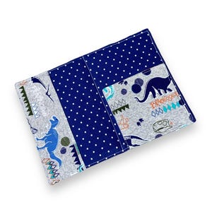 K&ouml;nnte beinhalten: Ein rechteckiger Stoffartikel mit einem Patchwork-Design. Das Design umfasst einen Abschnitt aus marineblauem Stoff mit wei&szlig;en Punkten und Abschnitte aus grauem Stoff mit blauen Dinosaurier-Illustrationen und dem Wort "Brachiosaur".