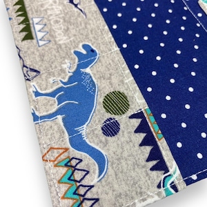 K&ouml;nnte beinhalten: Nahaufnahme eines Stoffartikels mit Dinosaurier-Motiv. Das Design zeigt einen blauen Dinosaurier, geometrische Formen und das Wort "Pteranodon" auf grauem Hintergrund. Ein Abschnitt aus blauem Stoff mit wei&szlig;en Punkten ist ebenfalls sichtbar.