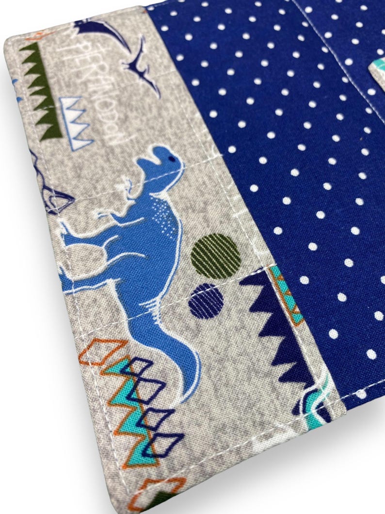 K&ouml;nnte beinhalten: Nahaufnahme eines Stoffartikels mit Dinosaurier-Motiv. Das Design zeigt einen blauen Dinosaurier, geometrische Formen und das Wort "Pteranodon" auf grauem Hintergrund. Ein Abschnitt aus blauem Stoff mit wei&szlig;en Punkten ist ebenfalls sichtbar.