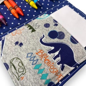 K&ouml;nnte beinhalten: Ein blau-grauer Stoff-Stifthalter mit Dinosaurier-Motiv. Der Halter hat eine Tasche f&uuml;r Buntstifte und eine separate Tasche f&uuml;r Papier. Der Stoff ist mit Dinosauriern, Rauten und Punkten gemustert. Buntstifte in verschiedenen Farben sind sichtbar.