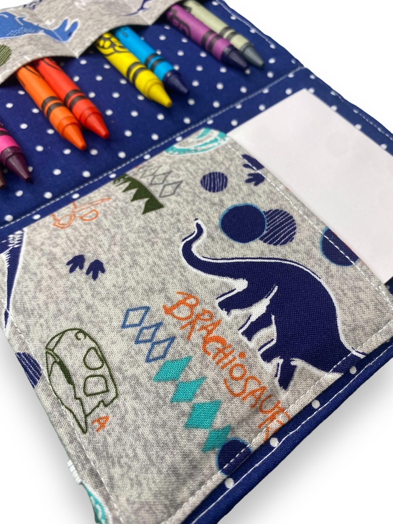 K&ouml;nnte beinhalten: Ein blau-grauer Stoff-Stifthalter mit Dinosaurier-Motiv. Der Halter hat eine Tasche f&uuml;r Buntstifte und eine separate Tasche f&uuml;r Papier. Der Stoff ist mit Dinosauriern, Rauten und Punkten gemustert. Buntstifte in verschiedenen Farben sind sichtbar.