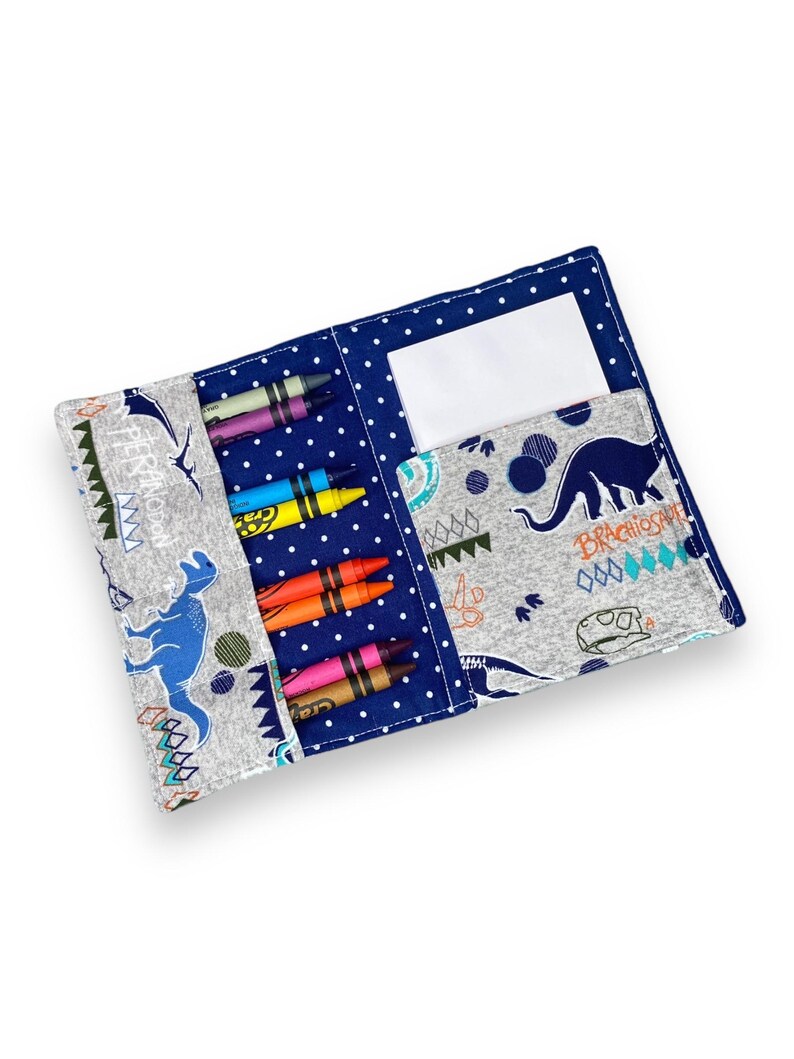 K&ouml;nnte beinhalten: Offenes Stiftetui mit blauem Tupfen- und Dinosaurier-Print-Design. Das Etui ist ge&ouml;ffnet und zeigt mehrere bunte Buntstifte und eine wei&szlig;e Karte. Das Etui ist aus Stoff und hat eine Tasche f&uuml;r die Buntstifte.
