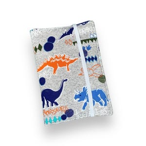 K&ouml;nnte beinhalten: Eine graue Stoff-Notizbuchh&uuml;lle mit Dinosaurier-Muster. Die H&uuml;lle zeigt blaue, orangefarbene und wei&szlig;e Dinosaurier-Illustrationen, geometrische Formen und das Wort "Pachiosaurus". Ein wei&szlig;es Gummiband sichert das Notizbuch.