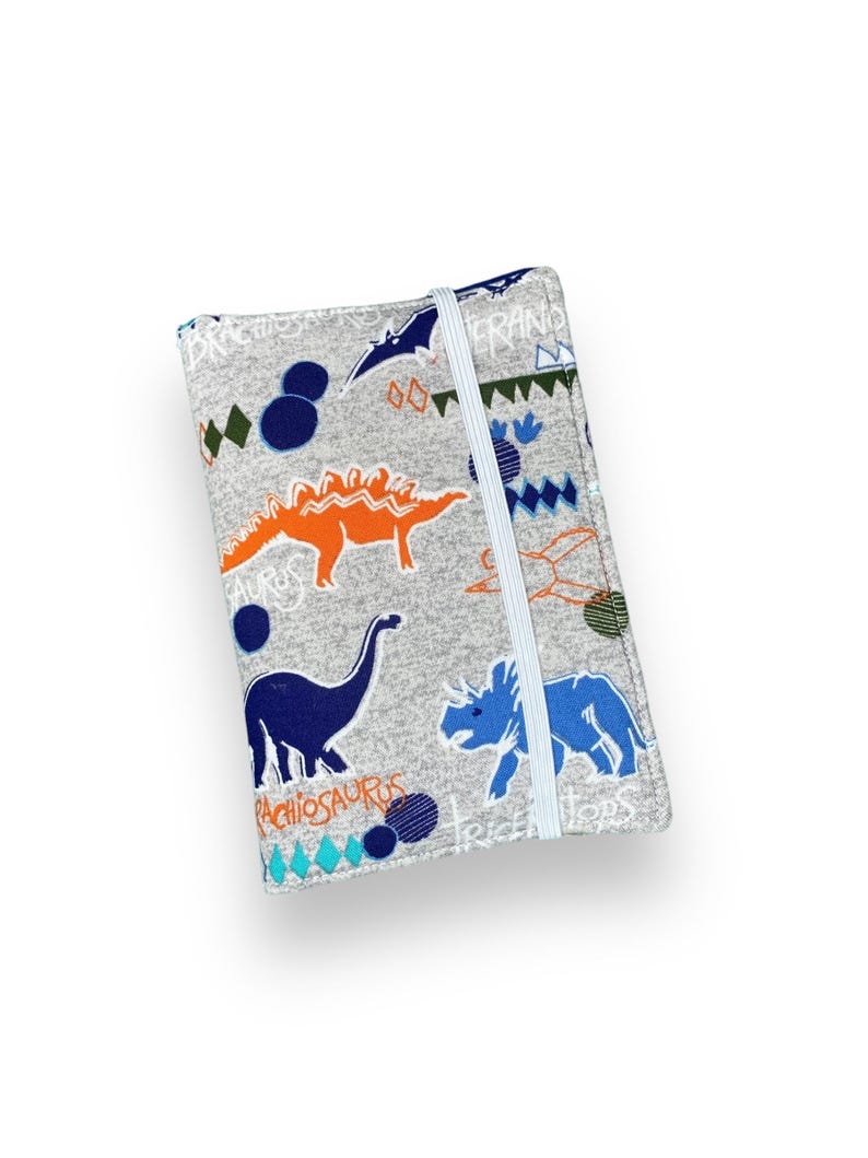 K&ouml;nnte beinhalten: Eine graue Stoff-Notizbuchh&uuml;lle mit Dinosaurier-Muster. Die H&uuml;lle zeigt blaue, orangefarbene und wei&szlig;e Dinosaurier-Illustrationen, geometrische Formen und das Wort "Pachiosaurus". Ein wei&szlig;es Gummiband sichert das Notizbuch.