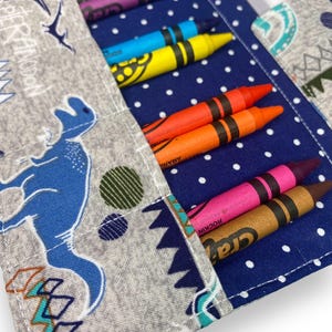 K&ouml;nnte beinhalten: Ein Stoff-Stifthalter mit blauem Tupfenmuster und grauem Dinosaurier-Print. Der Halter enth&auml;lt mehrere bunte Buntstifte, darunter lila, blau, gelb, orange, rosa und braun. Der Halter ist offen und zeigt die Stifte.