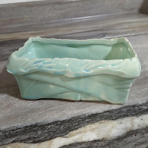 May include: A rectangular ceramic planter with a light mint green glaze and a textured, uneven rim. The exterior features a molded, overlapping leaf-like pattern that wraps around the sides, creating a dimensional and organic appearance.