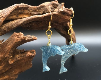 Könnte beinhalten: Ein Paar blaue Glitzer-Delfin-Ohrringe mit goldenen Haken. Die Delfine sind in entgegengesetzte Richtungen gerichtet und hängen an den Haken.