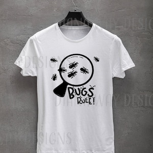 Puede incluir: Camiseta blanca con un dise&ntilde;o negro de una lupa llena de insectos. El texto "BUGS RULE!" est&aacute; debajo. La camiseta est&aacute; colgada en una percha negra sobre un fondo gris.