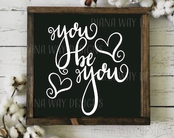 Op de afbeelding: Een houten bord met een zwarte achtergrond en witte cursieve tekst die "you be you" luidt met hartaccenten. Het bord is omgeven door een donkerbruine lijst en katoenstelen.