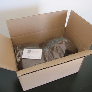 May include: A brown cardboard box with the top flap open, revealing two items wrapped in clear plastic. A handwritten note with the text "EWE&AG Thank you!" is visible on top of the items.