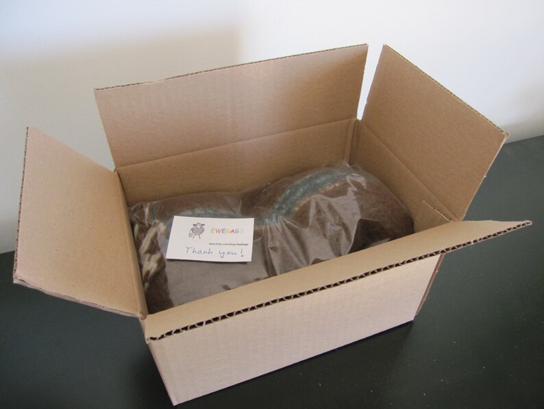 May include: A brown cardboard box with the top flap open, revealing two items wrapped in clear plastic. A handwritten note with the text "EWE&AG Thank you!" is visible on top of the items.