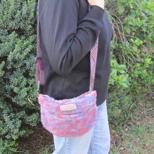 K&ouml;nnte beinhalten: Eine Person tr&auml;gt ein schwarzes Shirt mit Stehkragen und blaue Jeans und tr&auml;gt eine gestrickte Tasche in Lila und Rosa mit einem braunen Lederanh&auml;nger, auf dem "remarkable" steht.