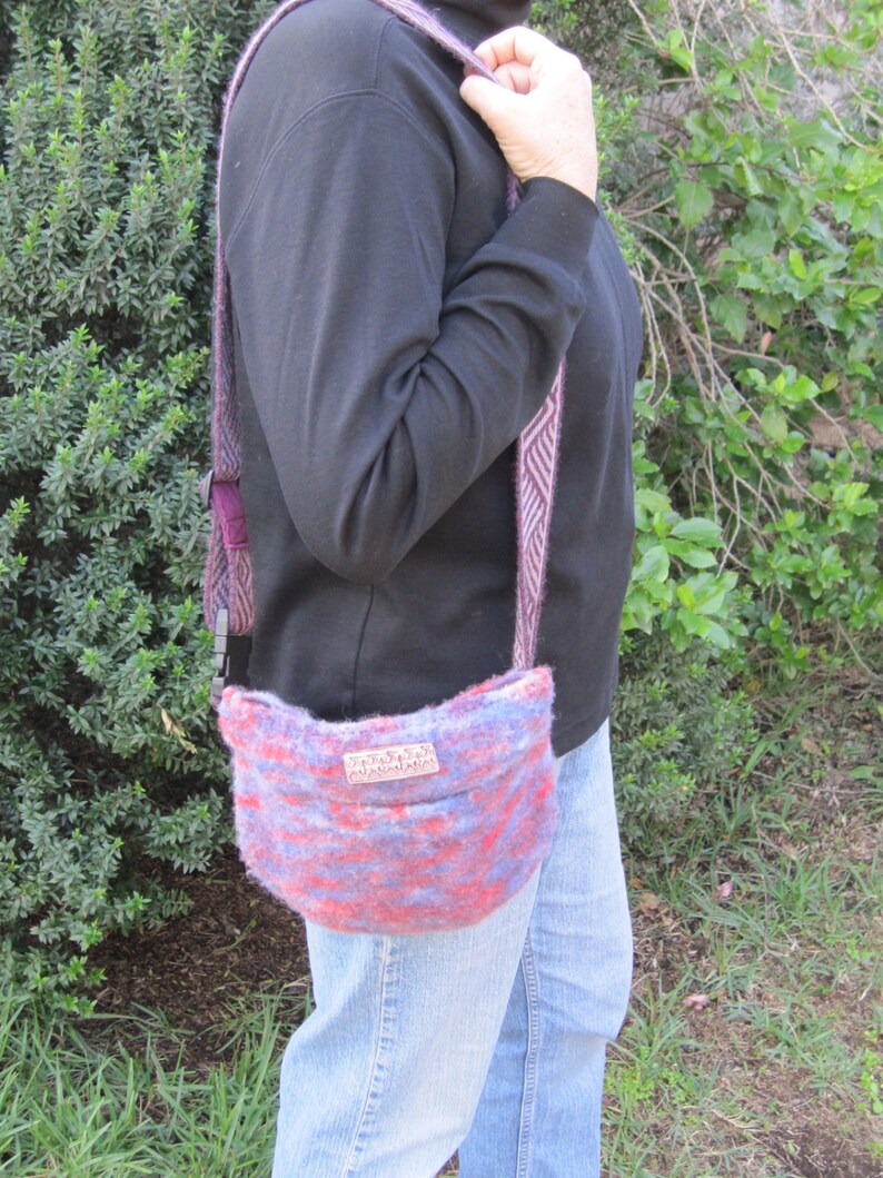 K&ouml;nnte beinhalten: Eine Person tr&auml;gt ein schwarzes Shirt mit Stehkragen und blaue Jeans und tr&auml;gt eine gestrickte Tasche in Lila und Rosa mit einem braunen Lederanh&auml;nger, auf dem "remarkable" steht.