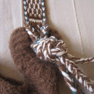 May include: A brown wool pouch with a white and blue braided strap. The strap is tied in a knot at the top of the pouch.