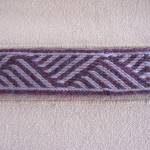 K&ouml;nnte beinhalten: Nahaufnahme eines gewebten Stoffbandes mit einem geometrischen Muster in Violett- und Graut&ouml;nen. Das Band ist auf einen beigefarbenen Stoffgrund gen&auml;ht.