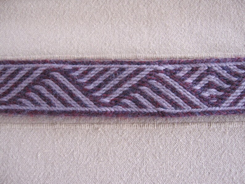 K&ouml;nnte beinhalten: Nahaufnahme eines gewebten Stoffbandes mit einem geometrischen Muster in Violett- und Graut&ouml;nen. Das Band ist auf einen beigefarbenen Stoffgrund gen&auml;ht.