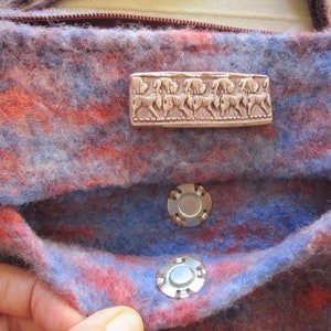 K&ouml;nnte beinhalten: Eine blaue und rote Wolltasche mit einem silbernen Metallverschluss und einem silbernen Metallobjekt mit einem Pferdemotiv. Die Tasche hat eine Klappe mit einem Druckknopfverschluss.