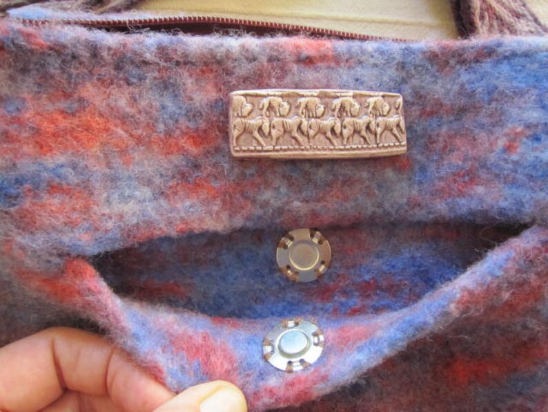 K&ouml;nnte beinhalten: Eine blaue und rote Wolltasche mit einem silbernen Metallverschluss und einem silbernen Metallobjekt mit einem Pferdemotiv. Die Tasche hat eine Klappe mit einem Druckknopfverschluss.