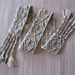 May include: A woven belt with a geometric diamond pattern in white, teal, and gold. The belt has braided tassels at the ends. The belt is lying on a textured, light brown surface.