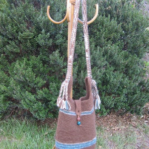 May include: A brown knitted tote bag with blue and white stripes and tassels. The bag is hanging on a wooden rack with a brown and white patterned strap.