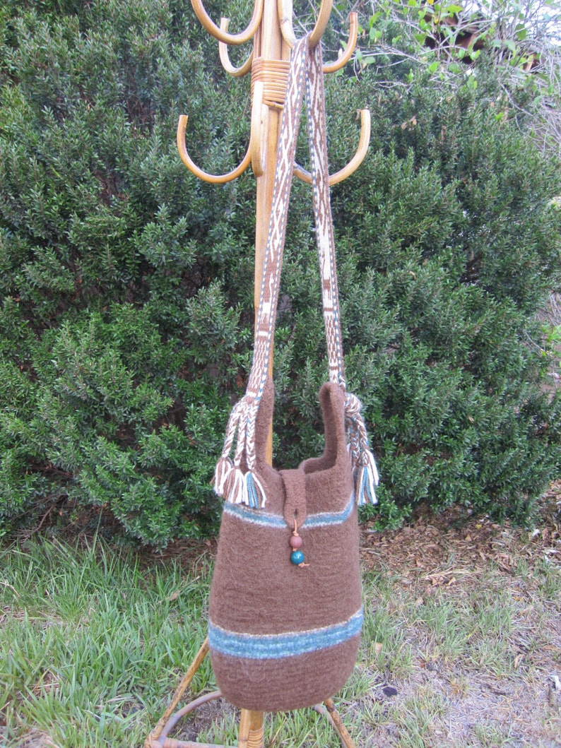 May include: A brown knitted tote bag with blue and white stripes and tassels. The bag is hanging on a wooden rack with a brown and white patterned strap.