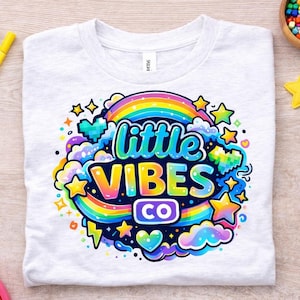 Puede incluir: Camiseta blanca con un diseño gráfico colorido que presenta un arcoíris, estrellas y las palabras "little VIBES CO." El diseño está rodeado de nubes y relámpagos. También se ven rotuladores de colores, un juguete de arcoíris y un cubo de Rubik.