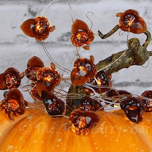 May include: A string of lights with acorn-shaped bulbs, wrapped around a pumpkin. The lights are a warm amber color and the pumpkin is orange.