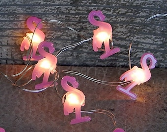 May include: A string of fairy lights with pink flamingo shaped bulbs. The lights are strung on a silver wire and are lit with warm white LED bulbs.