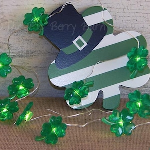 May include: A St. Patrick's Day themed string light with glowing green shamrocks and a leprechaun hat cutout. The hat is navy blue and white with a green stripe design. The lights are on a thin silver wire.
