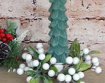 May include: A teal Christmas tree-shaped candle sits on a glass plate, surrounded by white berries and green leaves. A small arrangement of pine needles, red berries, and a pinecone is on the left. The background is a brick wall.