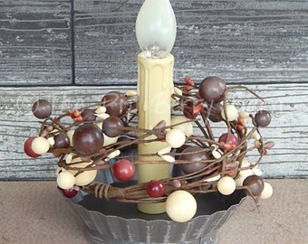 May include: A battery-operated candle with a cream-colored wax appearance and a flickering flame. The candle is surrounded by a decorative berry garland in shades of brown, cream, and red, set in a galvanized metal dish.