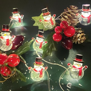 May include: String of clear snowman lights with red scarves and black top hats. The string is intertwined with red berries, green leaves, and a pinecone. The lights are on a dark green background.