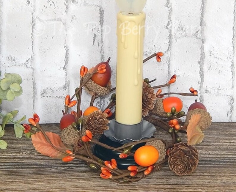 May include: A candle holder with a white pillar candle and a fall-themed arrangement of faux acorns and berries. The candle holder is made of metal and has a rustic finish.