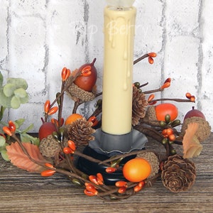 May include: A candle ring with a white pillar candle, brown branches, orange berries, brown acorns, and pine cones.