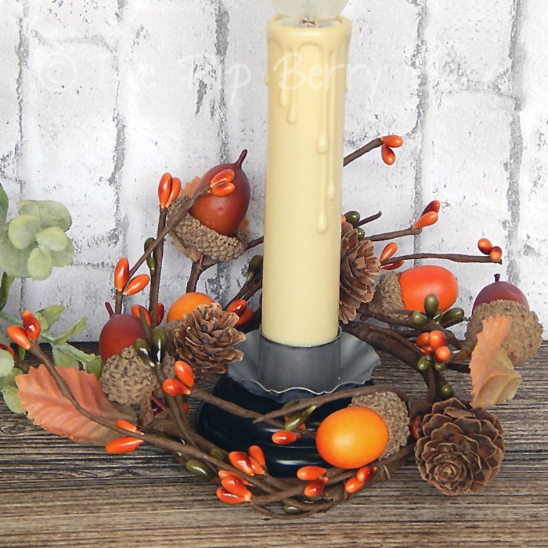 May include: A candle ring with a white pillar candle, brown branches, orange berries, brown acorns, and pine cones.