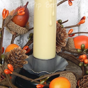 May include: A white pillar candle with a drip of wax sits in a gray metal candle holder. The candle is surrounded by a fall-themed wreath with brown acorns, orange berries, and pine cones.