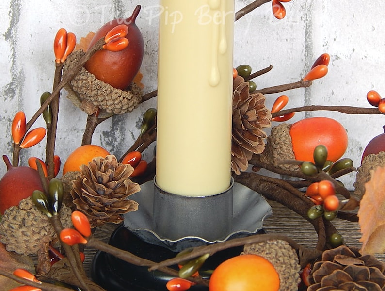 May include: A white pillar candle with a drip of wax sits in a gray metal candle holder. The candle is surrounded by a fall-themed wreath with brown acorns, orange berries, and pine cones.