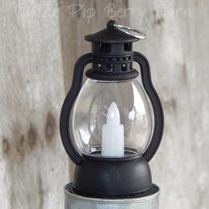 May include: A small black lantern with a clear, rounded glass enclosure. Inside is a white, battery-operated candle. The lantern has a handle and a ring at the top. The text "The Pip Berry Barn" is visible at the top of the image.