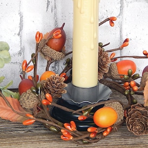 May include: A candle ring with a white candle, brown pine cones, orange and brown acorns, and orange berries. The candle ring is made of brown twigs and is sitting on a wooden surface.